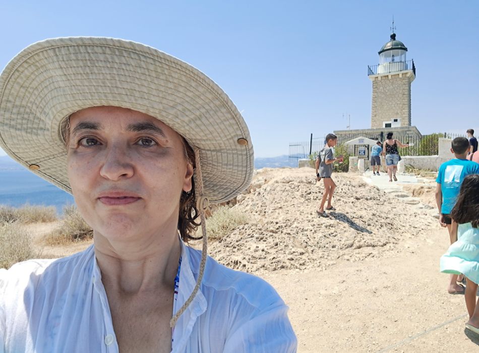 Evgenia, owner of TRavel the GReek way, wearing a hat, smiling in front of  Melagkavi Lighthouse in Loutraki