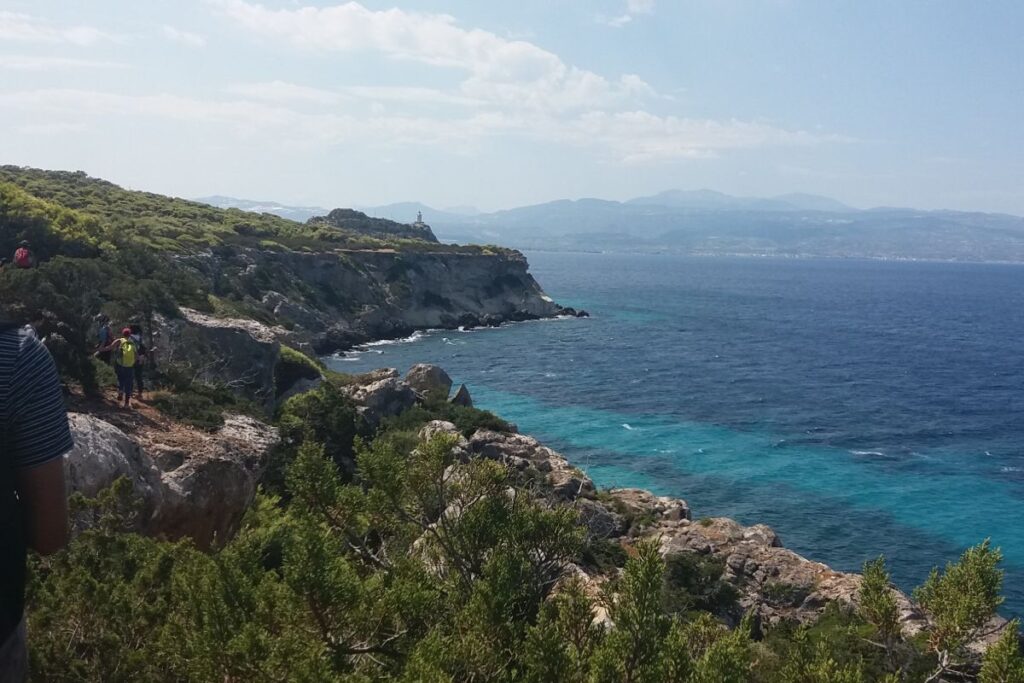 Hikers walking by a trail next to the sea with Melagkavi lighthouse in the background.