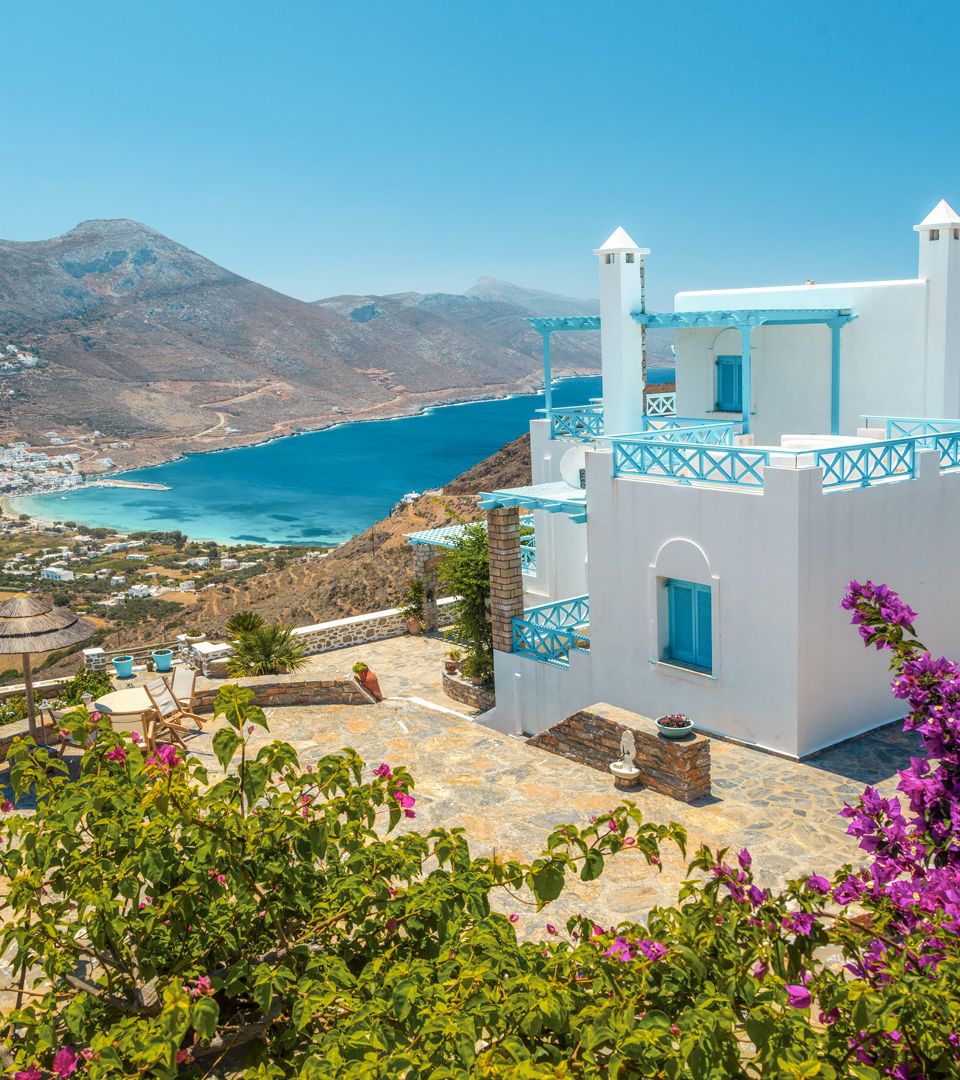A whitewashed house with blue windows with flowers and a large balcony having a view to the sea in a sunny day in Amorgos Island Greece.