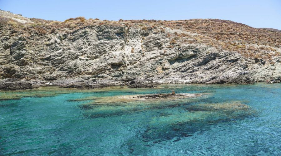 The image captures the serene waters of Livadaki Beach on Folegandros Island, Greece. The turquoise sea is crystal clear, revealing submerged rock formations beneath the surface. The rocky shoreline rises steeply from the water, covered with dry, sparse vegetation typical of a Mediterranean climate. The combination of vibrant blue waters and rugged, rocky landscape creates a picturesque coastal scene, showcasing the natural beauty and tranquility of this remote beach location.