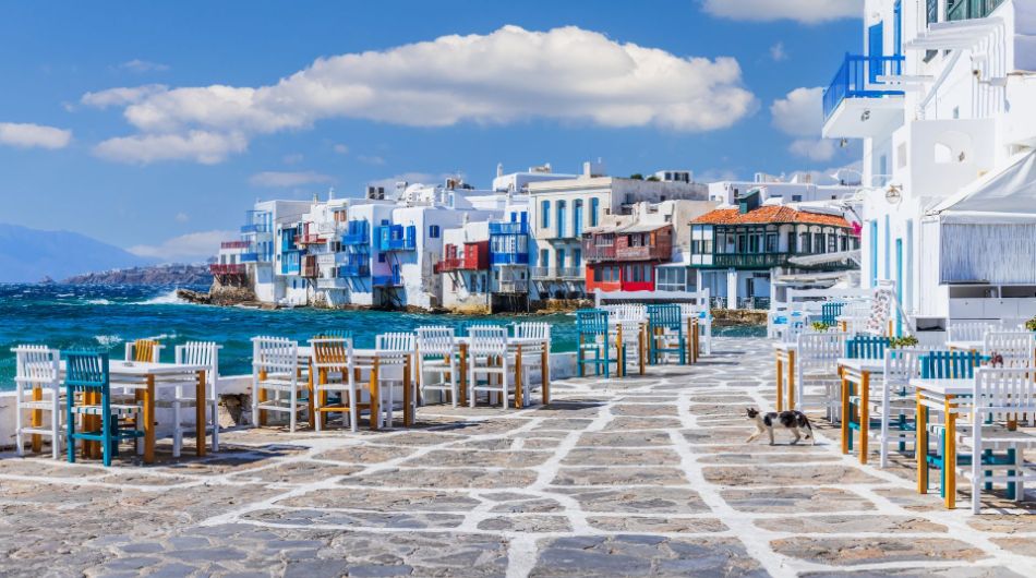 Tables and chairs by the sea of Little Venice in Mykonos with a cat walking and colorful houses  in the background.
In Little Venice in Mykonos.