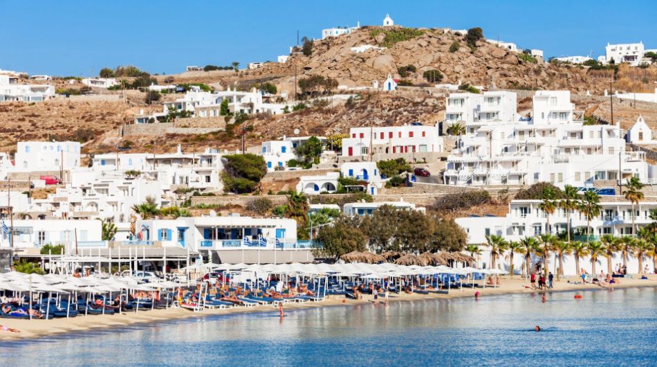 Ornos Beach Santa Marina with many people by the sea under umbrellas.
In the background whitewashed houses.
In Mykonos Island.
