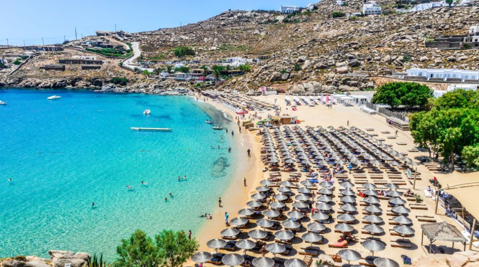 Many people sitting be the sea under umbrellas. Trees in the background and some whitewashed houses.
In Super Paradise Beach in Mykonos Island. 