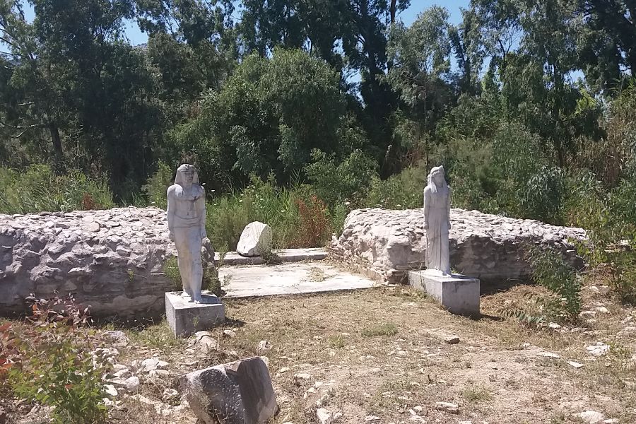 Two ancient statues stand at the Sanctuary of the Egyptian Gods in Marathon, Greece, surrounded by greenery and remnants of old stone structures under a sunny sky. 