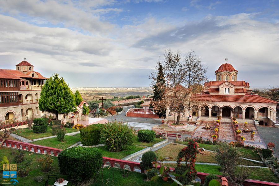 The picturesque Agios Efrem Monastery in Marathon, Greece, featuring beautifully landscaped gardens, traditional architecture with red-tiled roofs, and a scenic view of the surrounding landscape under a partly cloudy sky.