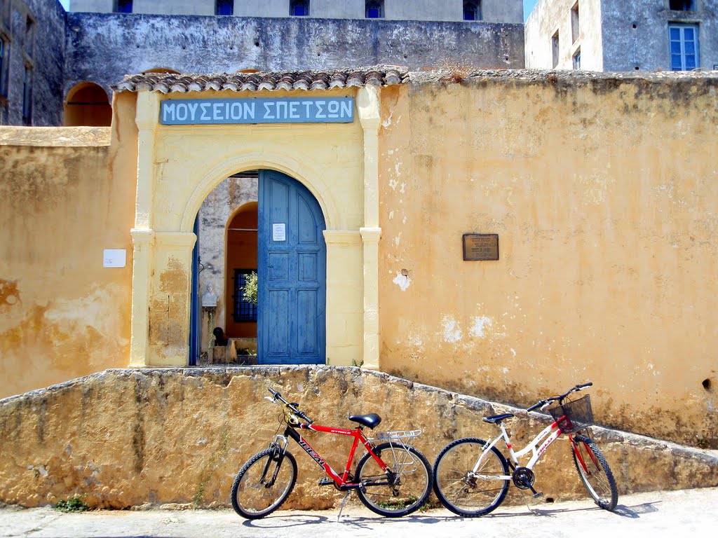 "The entrance to the Spetses Museum, featuring an arched doorway with a blue wooden door and a sign in Greek above it. The entrance is set in a weathered, ochre-colored stone wall. Two bicycles, one red and one white, are parked in front of the wall. The surrounding buildings have a rustic, aged appearance, contributing to the historic charm of the location."