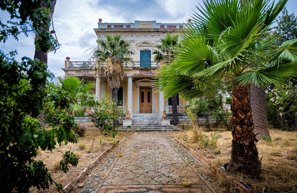 "A grand, historic mansion surrounded by lush palm trees and overgrown vegetation. The two-story building features a neoclassical design with a columned entrance, ornate balconies, and detailed architectural elements. A stone pathway leads up to the wooden front doors, flanked by statues and an intricate mosaic pattern on the ground. The scene evokes a sense of faded opulence and timeless elegance under a cloudy sky."