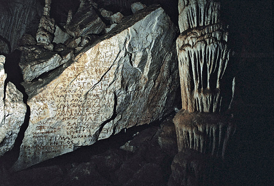 Interior of Chrysospilia Cave on Folegandros Island, Greece, featuring ancient inscriptions carved into a large rock surface. The dimly lit cave reveals stalactites and stalagmites with intricate formations around the engraved rock. The inscriptions, written in Greek, provide a glimpse into the cave’s historical significance and the mysterious atmosphere of the underground chamber.






