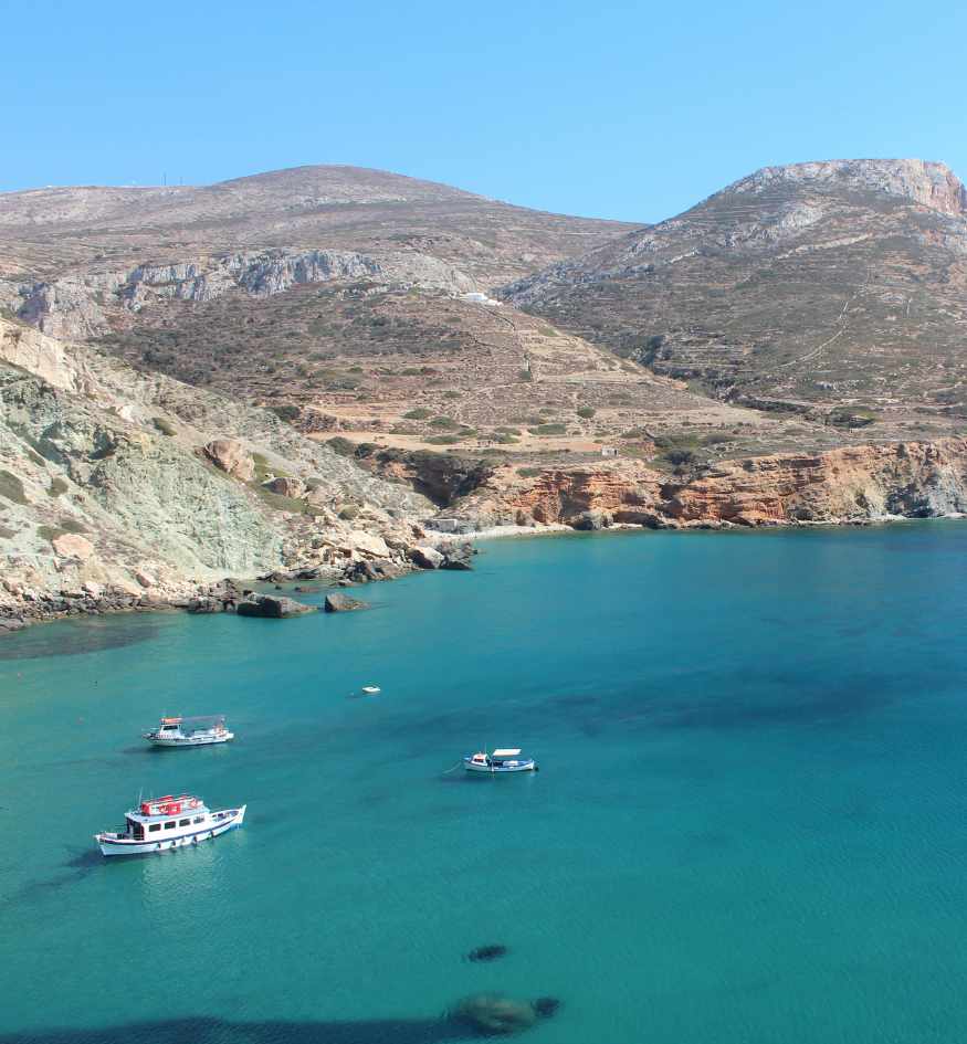 Turquoise sea with small boats below the rugged rocky hills and coastline of Folegandros, Greece.