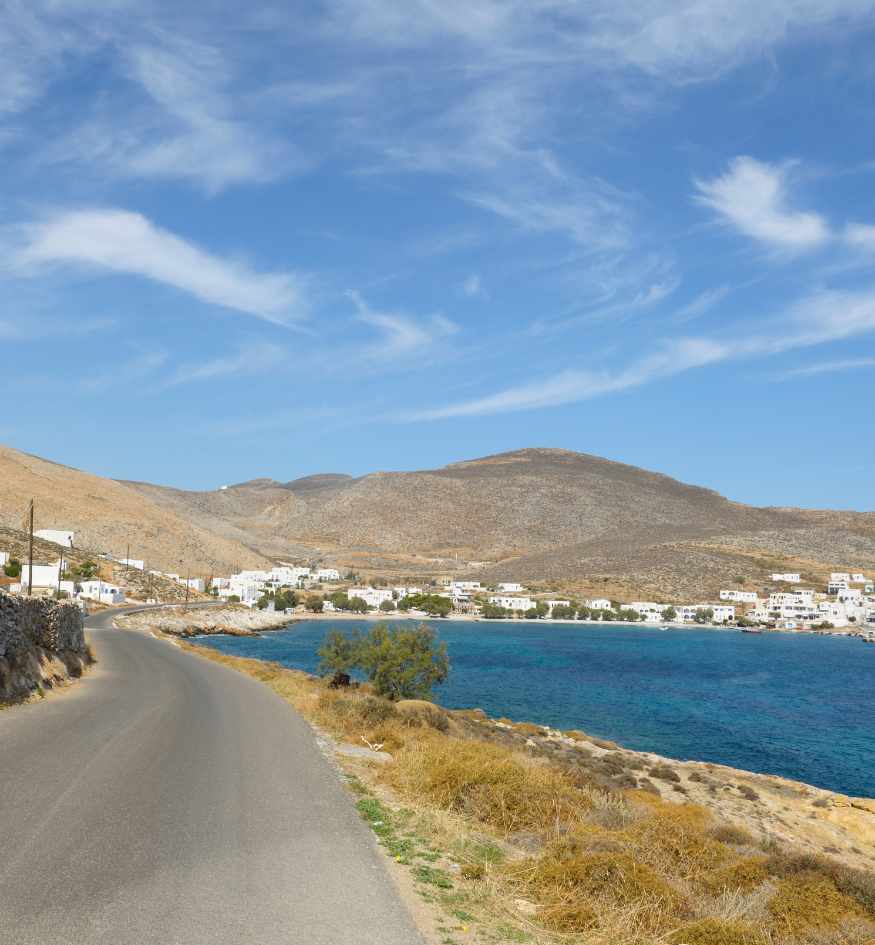 Quiet coastal road following a blue bay on Folegandros, Greece, with whitewashed houses and dry rocky hills in the distance.