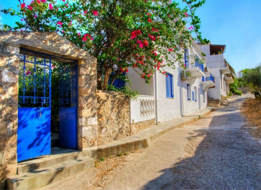 This image depicts a quaint, sunlit street on Spetses Island. The scene features traditional whitewashed houses with vibrant blue shutters and balconies, some adorned with wrought iron railings. A beautiful bougainvillea vine with bright pink flowers climbs over a stone archway entrance with blue metal gates, casting dappled shadows on the stone wall. The narrow, slightly inclined street is paved and flanked by rustic buildings, leading into the distance under a clear, blue sky. The overall atmosphere is warm and inviting, showcasing the charm and picturesque nature of this Mediterranean village.