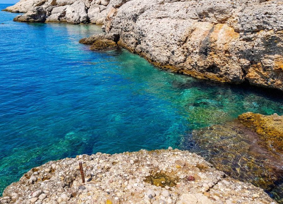 "Clear, turquoise waters of a rocky coastal area, with sunlight illuminating the underwater features and creating a gradient from deep blue to emerald green. The rocky shoreline is characterized by rugged, weathered rocks with visible strata and pebble-covered surfaces. The tranquil water contrasts with the rough texture of the rocks, creating a striking natural scene."