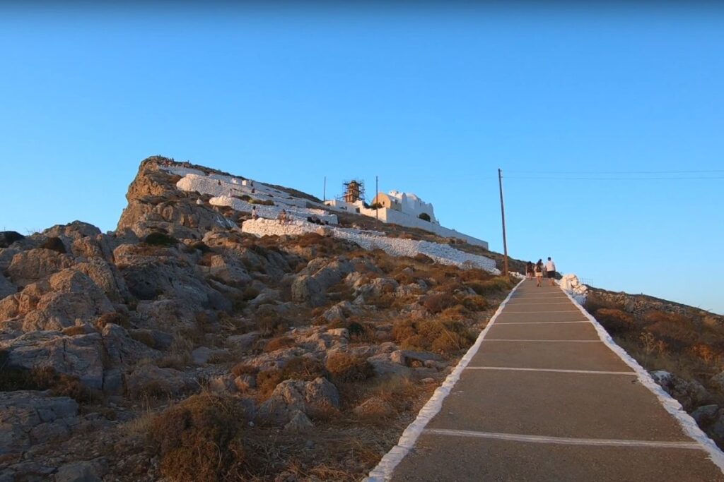 A path leading up to the Church of Panagia on Folegandros Island, Greece, at sunset. The path is lined with white stones and ascends a rocky hillside toward the white-washed church perched on top. The surrounding landscape features rugged terrain and sparse vegetation, illuminated by the warm glow of the setting sun. People walk along the path, adding a sense of pilgrimage and serenity to the scene against a backdrop of a clear blue sky transitioning to dusk.