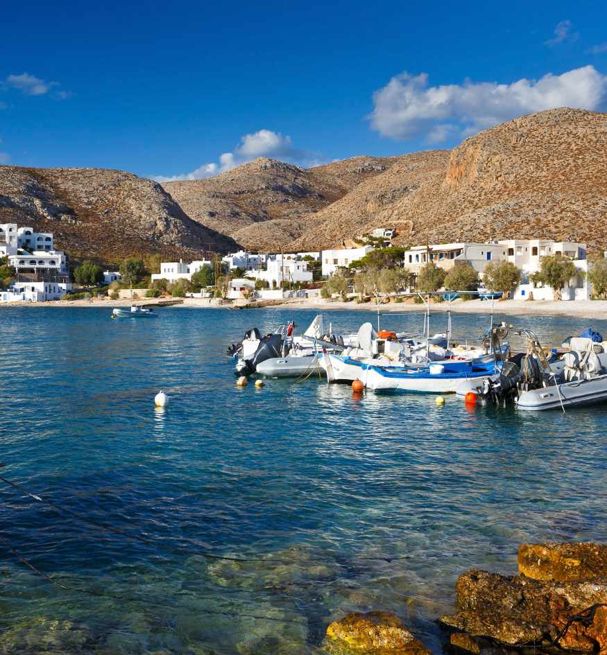 Small fishing boats in the clear blue harbor at Karavostasis, Folegandros, with whitewashed houses and rocky hills behind.