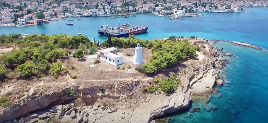 "An aerial view of a picturesque coastal town with a lighthouse on a rocky promontory surrounded by greenery. The lighthouse is white with an attached building and overlooks the turquoise waters of the bay. Several boats, including a large ship, are anchored in the calm harbor. The town in the background has charming white buildings with terracotta roofs, extending along the waterfront. The scene captures a blend of natural beauty and quaint maritime architecture."






