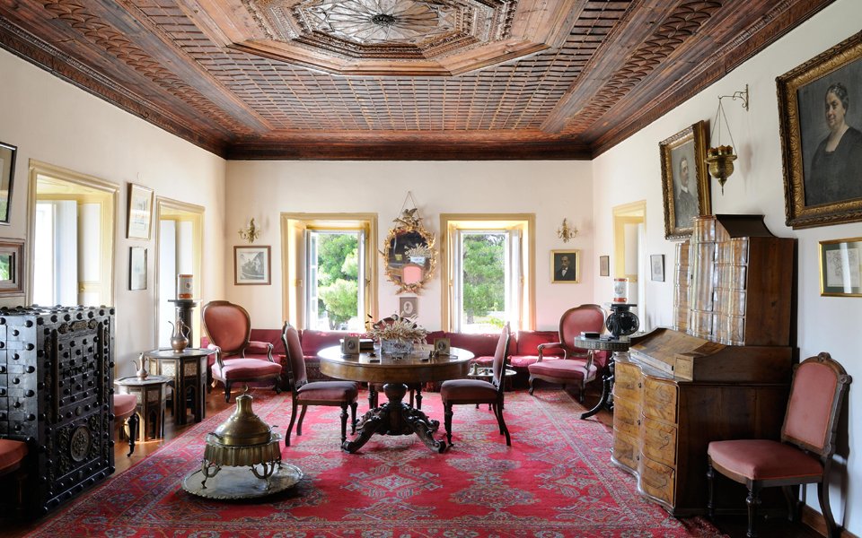 "An elegant, vintage room featuring ornate wooden furniture and a richly detailed wooden ceiling. The room is decorated with antique pieces, including a central round table surrounded by upholstered chairs, a large wooden cabinet, and a desk. Portraits and framed artworks adorn the walls, while large windows with yellow frames allow natural light to fill the space. The floor is covered with a patterned red carpet, and various decorative objects, such as lamps and vases, are displayed throughout the room. The setting reflects a sophisticated, historical ambiance."