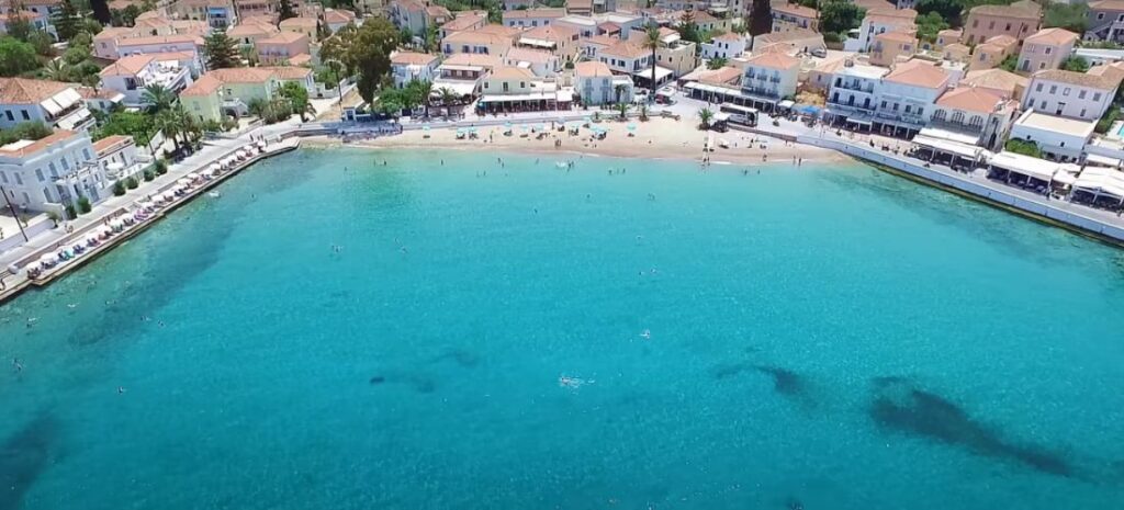 "An aerial view of a charming coastal town with a sandy beach curving around a clear, turquoise bay. The beach is lined with sunbathers and umbrellas, while swimmers enjoy the water. The town features traditional Mediterranean architecture, with white-washed buildings and terracotta roofs extending along the shoreline. A promenade with cafes and shops runs parallel to the beach, creating a lively waterfront atmosphere under a sunny sky."