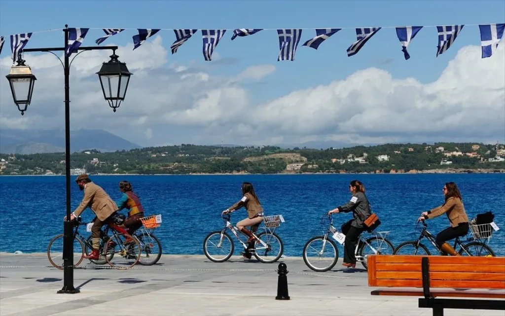 "A group of cyclists riding along a waterfront promenade under string flags with Greek patterns. The cyclists are dressed in vintage-style clothing, and their bicycles have baskets. The background features a vibrant blue sea, with distant hills and houses visible across the water. Traditional street lamps and a wooden bench are positioned along the promenade, creating a lively and scenic atmosphere on a clear day."