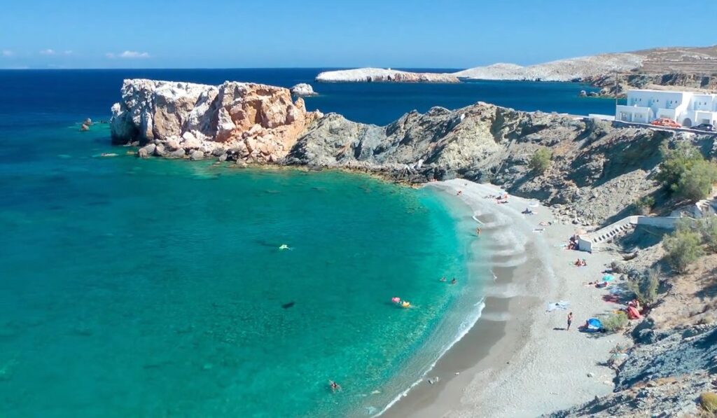 Aerial view of Vardia Beach on Folegandros Island, Greece, featuring a crescent-shaped sandy beach with turquoise waters. The beach is bordered by rugged cliffs and rocky formations, with a few white-washed buildings perched above. Sunbathers and swimmers enjoy the clear sea, while the bright blue sky enhances the serene coastal landscape. The scene captures the natural beauty and tranquil atmosphere of the secluded beach.