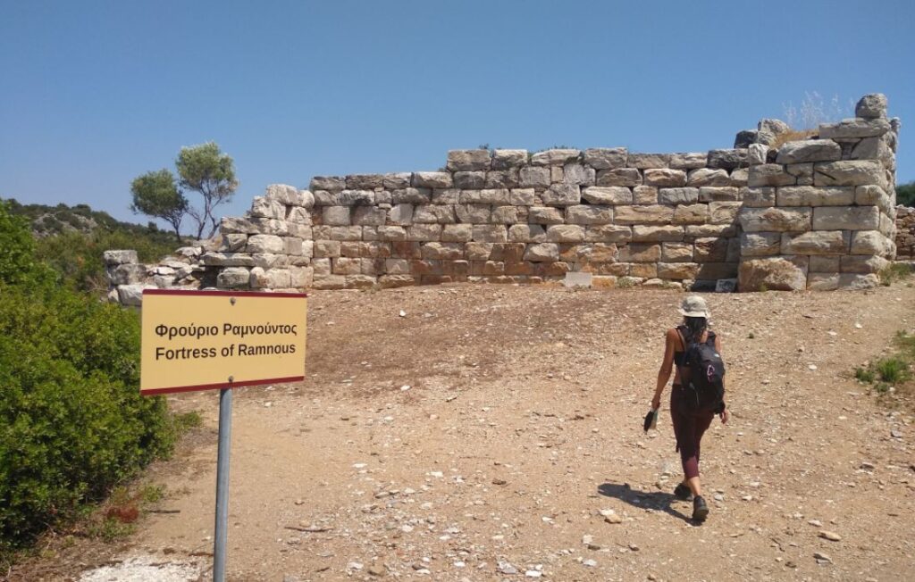 A hiker approaching the ruins of the Fortress of Rhamnous in Marathon, Greece, with a sign indicating the historical site, under a bright sky.