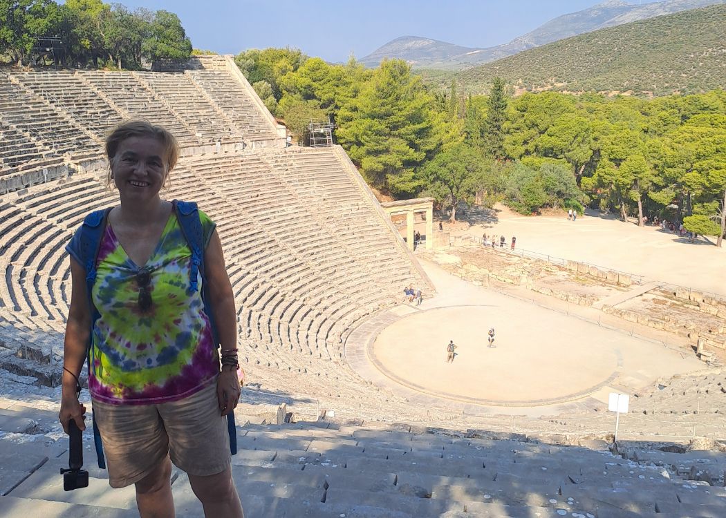  at the Epidaurus ancient theater in Greece, smiling with the expansive stone amphitheater and hills in the background.