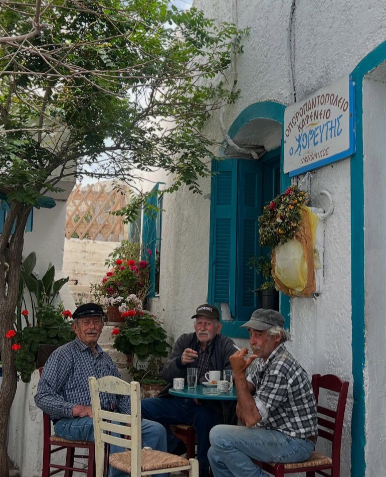 Three elderly men sitting at a small blue table outside a traditional whitewashed cafe with blue shutters, Amorgos, Greece.