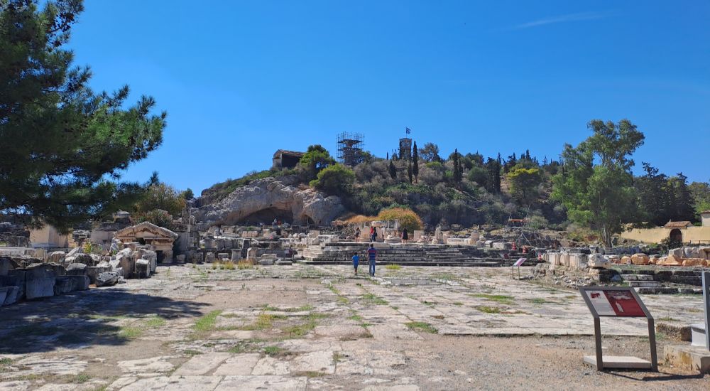 Ancient ruins of Eleusis archaeological site with stone pathways, steps, and scattered remains, surrounded by greenery and trees.