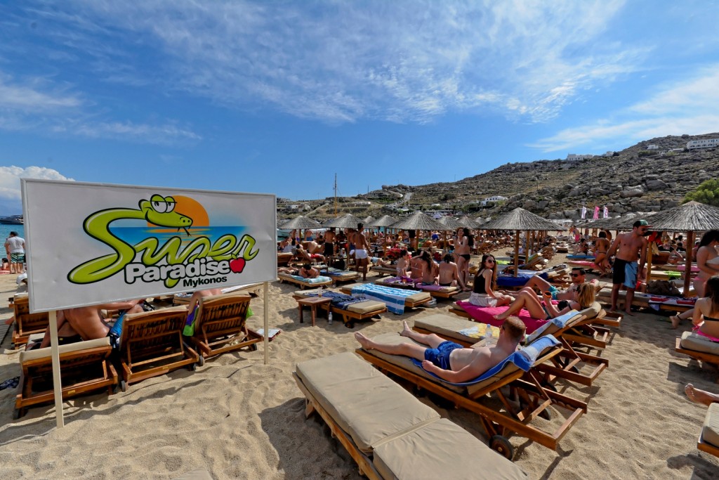 Many people sitting on the sandy beach under umbrellas in Super Paradise in Mykonos Island.