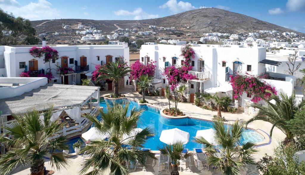 Aerial view of a charming Mediterranean hotel complex on Folegandros Island. The property features traditional whitewashed buildings adorned with vibrant bougainvillea and dark blue shutters. A central azure swimming pool is surrounded by lush palm trees and cozy shaded seating areas, offering a relaxing oasis. The picturesque setting is framed by rolling hills and whitewashed village houses dotting the landscape, providing a tranquil and authentic Greek island ambiance.