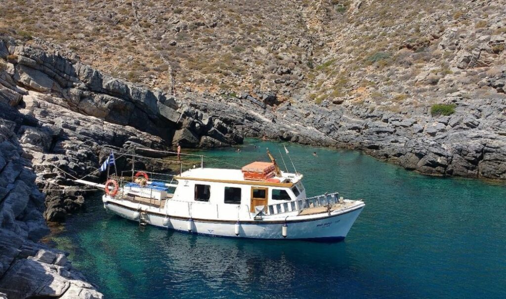 "A small, white motorboat anchored in a secluded, rocky cove with clear turquoise water. The boat has a covered cabin and is equipped with life rings and a Greek flag. The surrounding landscape is rugged and arid, with steep rocky cliffs leading down to the water."