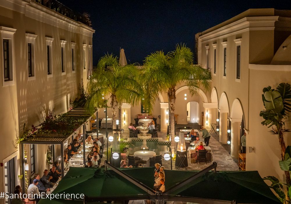 A vibrant evening dining scene in a beautiful courtyard with warm lighting and lush greenery in Santorini.