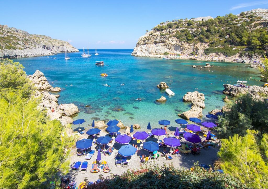A rocky beach called Anthony Quinns Beach with many blue umbrellas and boats in the sea in Rhodes Island.
