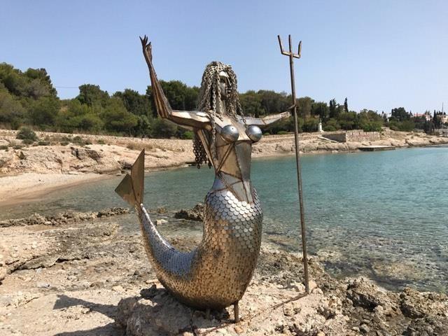 "A metallic mermaid statue stands on a rocky shoreline, overlooking calm turquoise waters. The sculpture depicts a mermaid with a scaled tail and outstretched arms, holding a trident in one hand. The mermaid has long, flowing hair made of metal strands. Behind her, a tree-lined shore extends along the water's edge under a clear sky. The scene combines art and nature in a coastal setting."