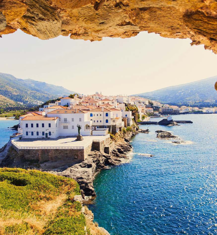 A high-angle, framed view through a rocky cave opening looking out at Chora, the capital of Andros, Greece. The town features white neoclassical buildings with red-tiled roofs situated on a narrow peninsula extending into the turquoise Aegean Sea. A statue stands in a wide stone plaza at the peninsula's tip.