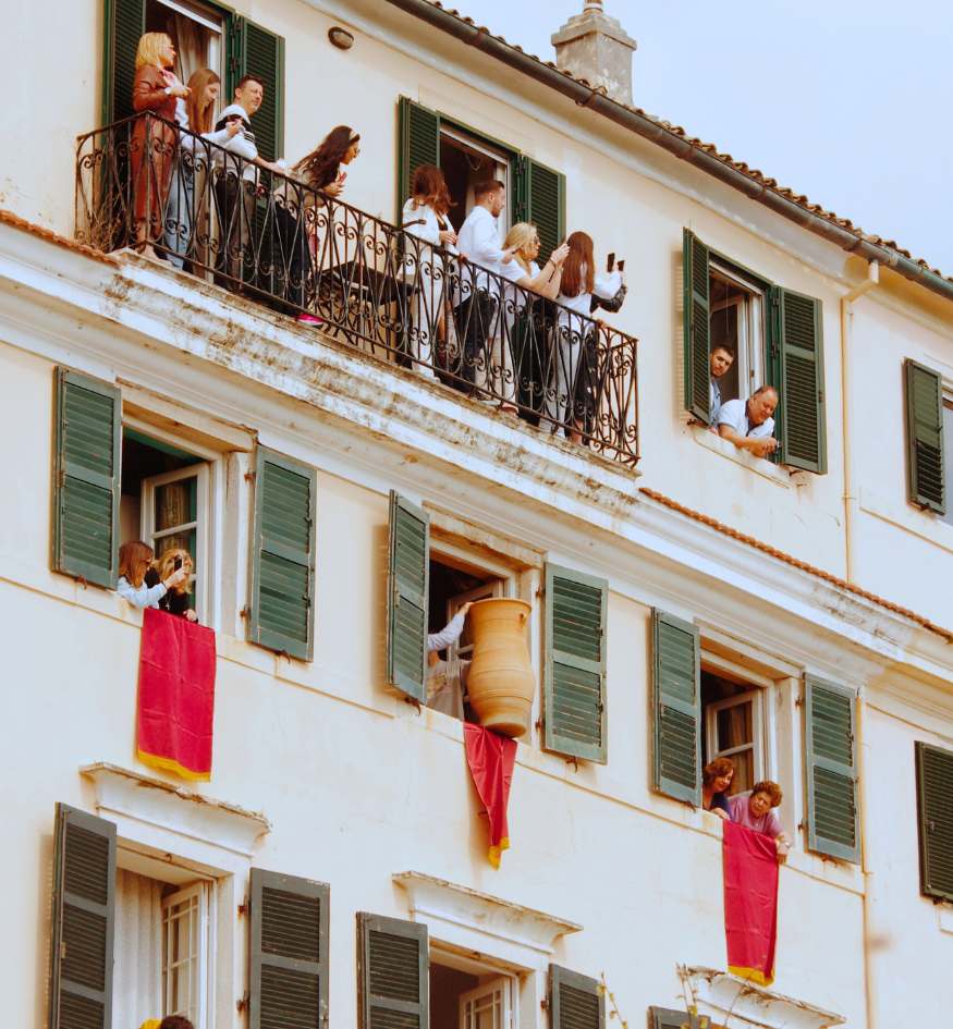 A multi-story white building in Corfu, Greece, during the "Botides" Easter tradition. Numerous people are leaning out of windows and over green-shuttered balconies. In the center, a person is mid-action, throwing a large clay pot (botis) draped with red ribbons from a window to the street below.