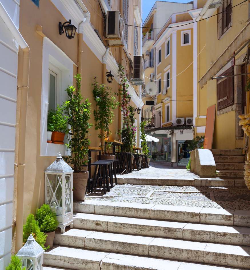 A bright, narrow pedestrian street in Corfu Town, Greece. The alley features whitewashed stone steps leading upward, lined with light yellow buildings. Outdoor seating with black stools and small green trees in pots are arranged outside a shop, and white decorative lanterns sit on the steps.