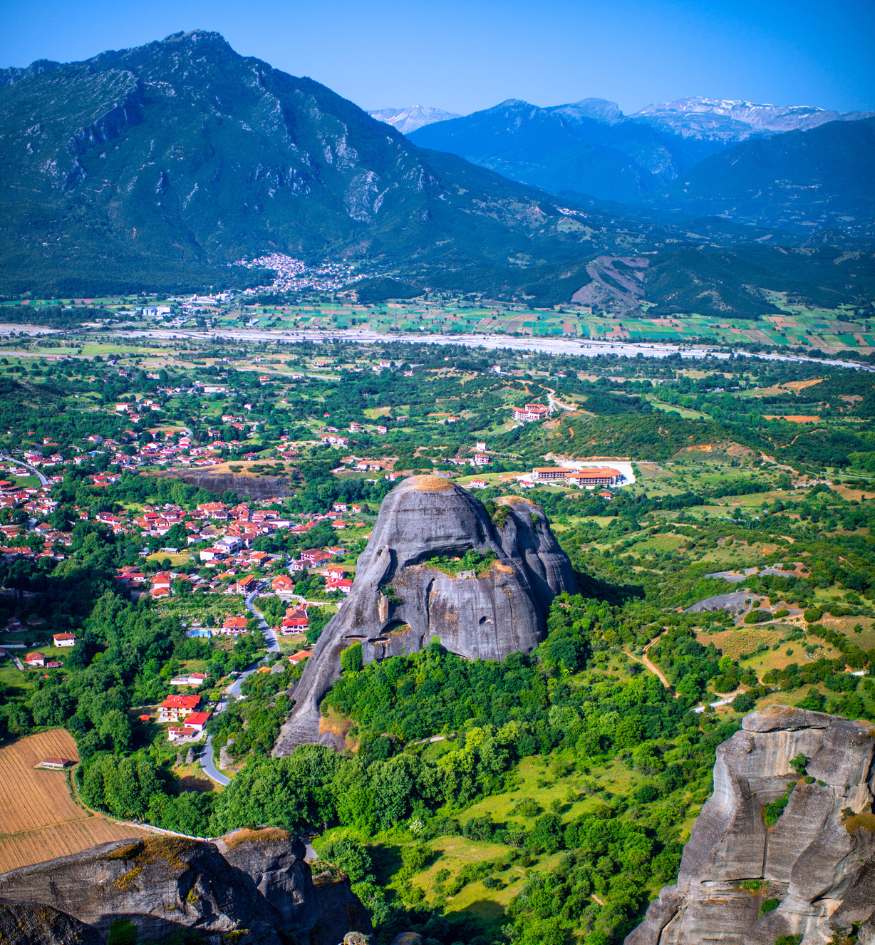 A wide, high-altitude landscape view of the Meteora region in Greece. In the foreground, a massive, rounded grey rock pillar is surrounded by vibrant green forests. The town of Kastraki with its red-roofed houses sits at the base. Tall, blue-toned mountains with snow-capped peaks are visible in the far distance.