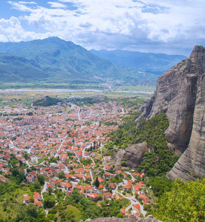 A high-angle view looking down over the town of Kalabaka, Greece. The town is a sea of red-tiled roofs nestled tightly against the base of towering, vertical dark grey conglomerate rock cliffs. A riverbed and green fields stretch out toward the hazy mountains in the background.