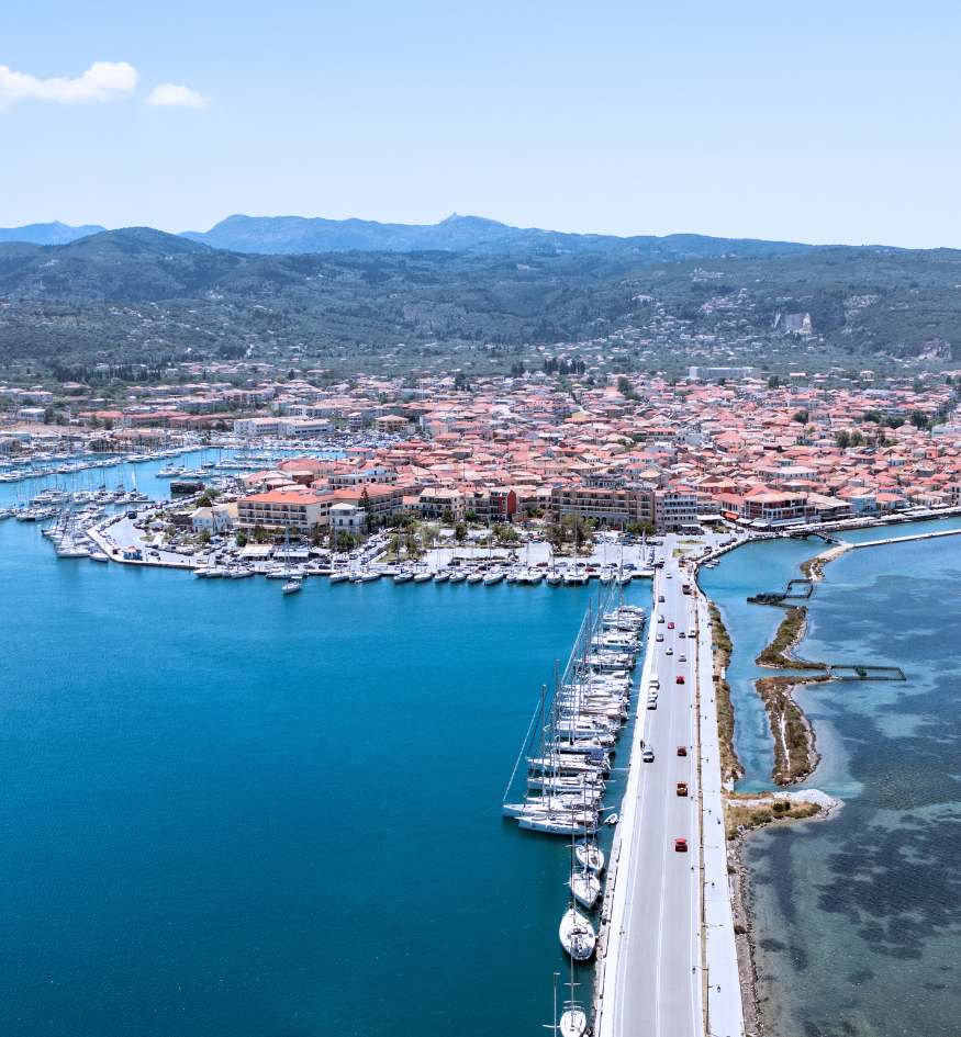 An aerial view of the entrance to Lefkada Town, Greece. A long, straight bridge filled with cars stretches across a bright blue lagoon toward the town. To the left, a large marina is packed with white sailboats and yachts. Lush green mountains rise in the distance under a clear sky.
