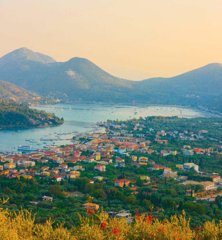High-angle view of Nydri village on Lefkada, showing a coastal town nestled between lush green hills and a bay filled with sailboats.