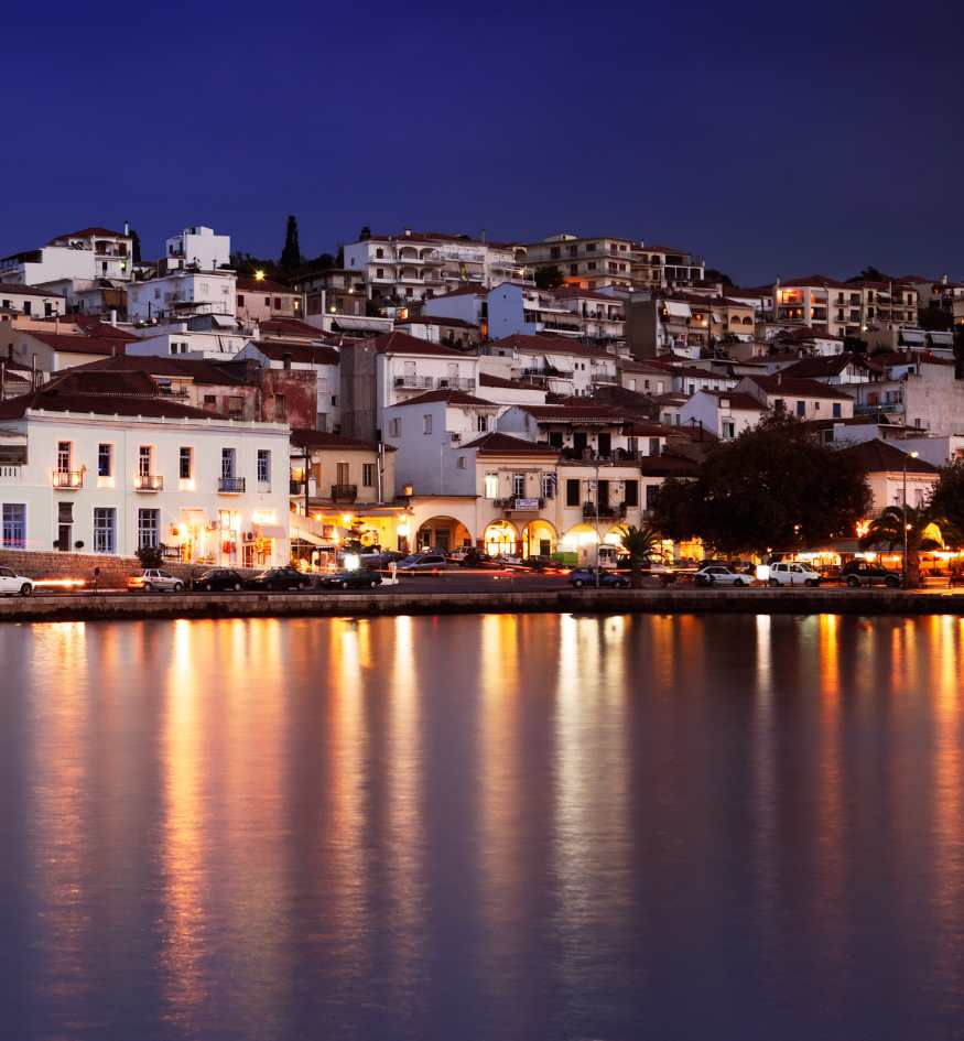 A nighttime view of Pylos, Greece, showing lit buildings and streetlights reflecting off the calm dark waters of the harbor.