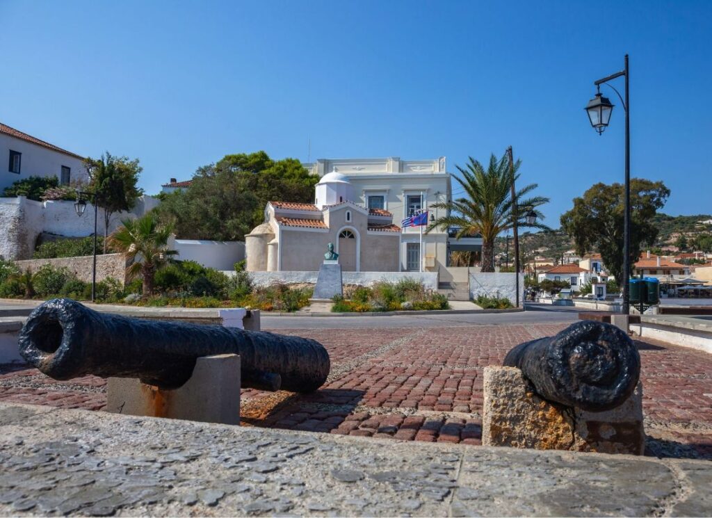 "A historical square featuring two old cannons positioned on either side of a cobblestone path. In the background, there is a large, white building with Greek flags, palm trees, and a small white chapel with a tiled roof. The area is surrounded by lush greenery, including shrubs and trees, under a clear blue sky. The scene captures a mix of historical and architectural elements in a Mediterranean setting."