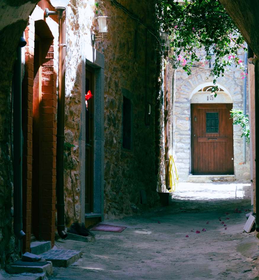 A sun-drenched, narrow stone alleyway in a Mesta village, Chios, Greece. The medieval architecture features heavy stone walls and arched doorways. A dark wooden door with a small window is visible at the end of the path, framed by shadows and lush green vines with pink flowers.