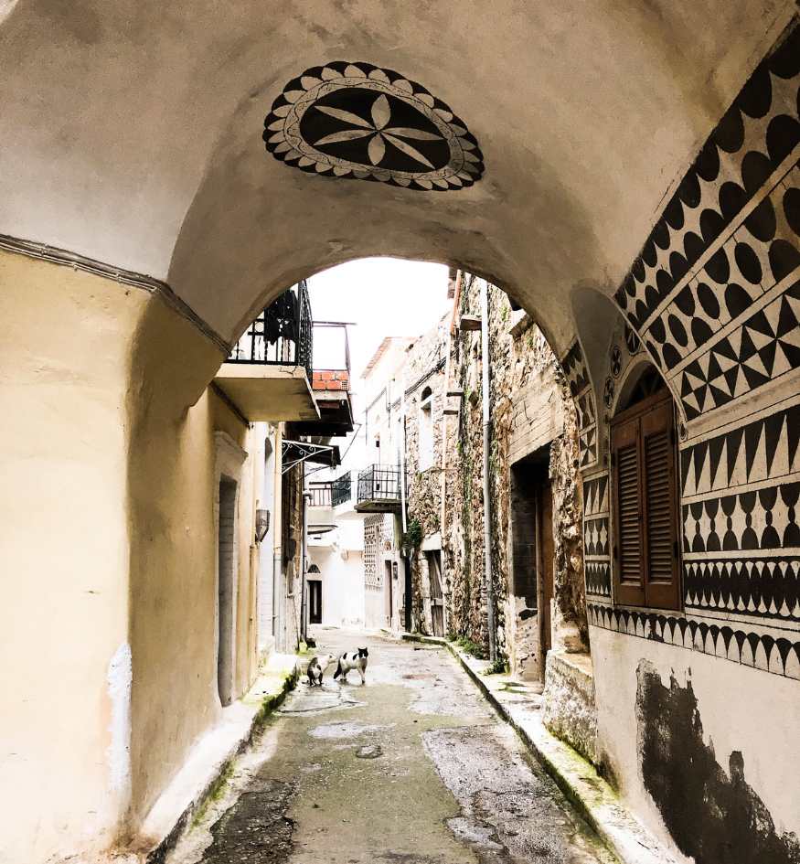 A narrow, paved alleyway in Pyrgi, Chios, Greece, seen through a stone archway. The building facades are decorated with "xysta"—intricate black and white geometric patterns. Two small cats stand in the middle of the wet street, and a decorative floral motif is painted on the underside of the arch.