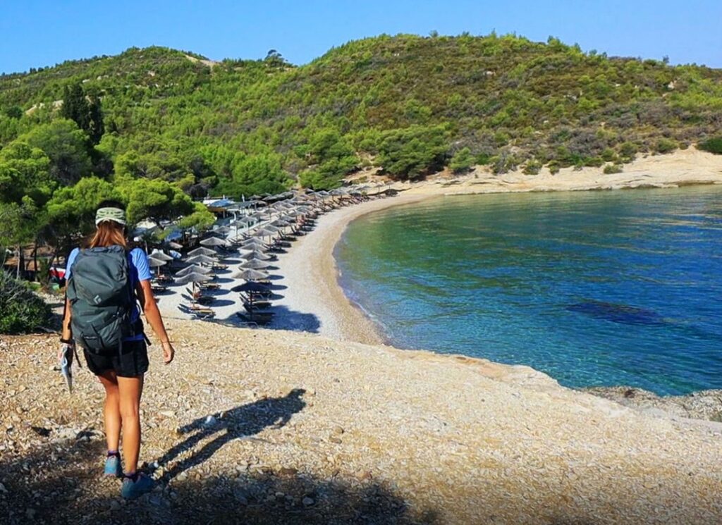 A sandy beach with many umbrellas and tree and a hiker on the cliff in Spetses Greece.  