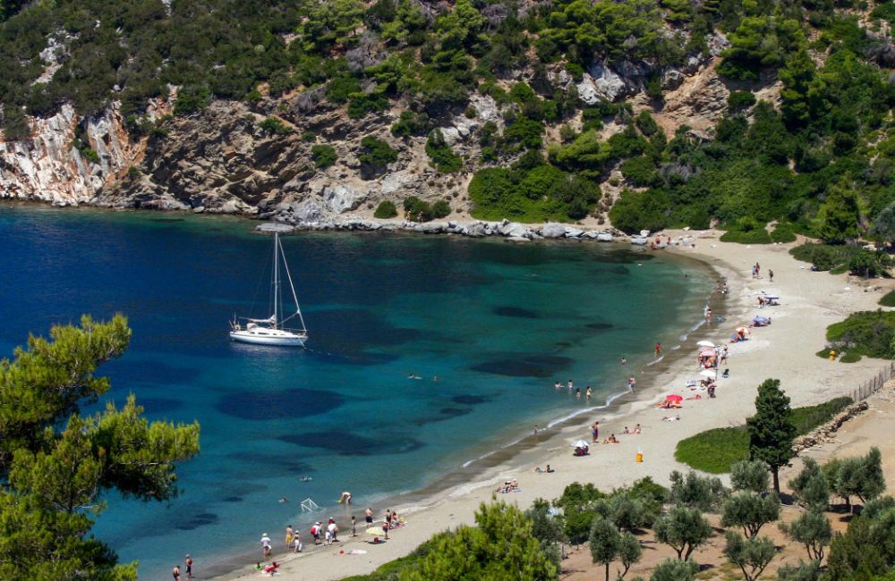 A sandy beach with many people, trees and bushes and a yacht in the sea in a sunny day. 