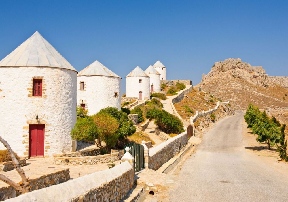 Five Windmills in Leros Island, a road and some trees in a sunny day. Off-The-Beaten-Track Greece Destinations.