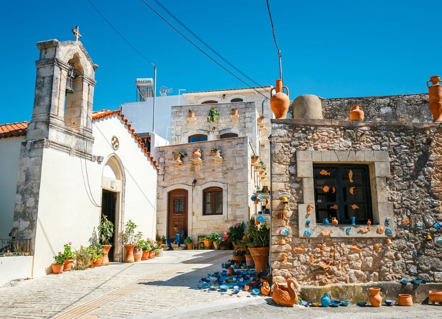 Charming courtyard in Margarites village, Rethymno, Crete, featuring traditional stone buildings adorned with terracotta pots and decorative pottery, with a small bell tower on the left.