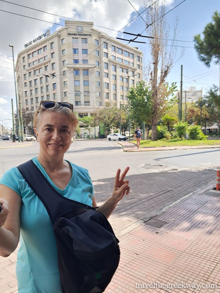 10 must-have items when you travel to Greece  and Evgenia wearing a cross body bag smiling in front of a hotel in Athens. 