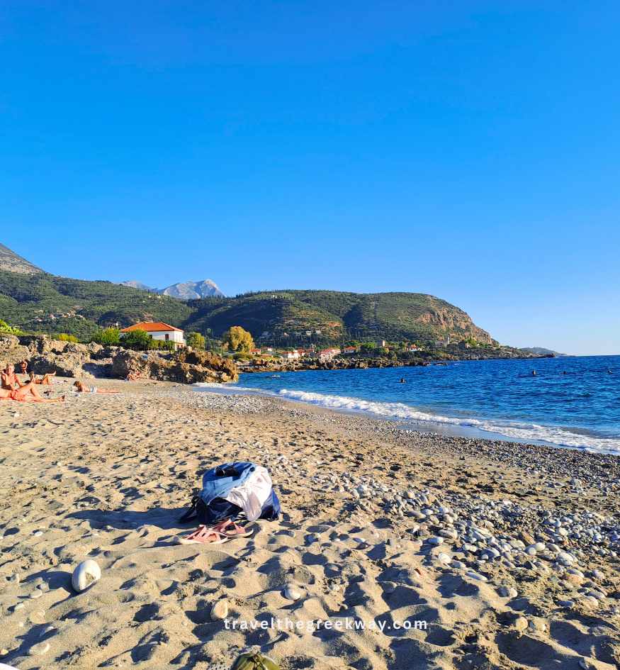 Sunny view of a pebble and sand beach in Mani, Greece, with clear blue water, stone houses, and rugged green hills in the background.
